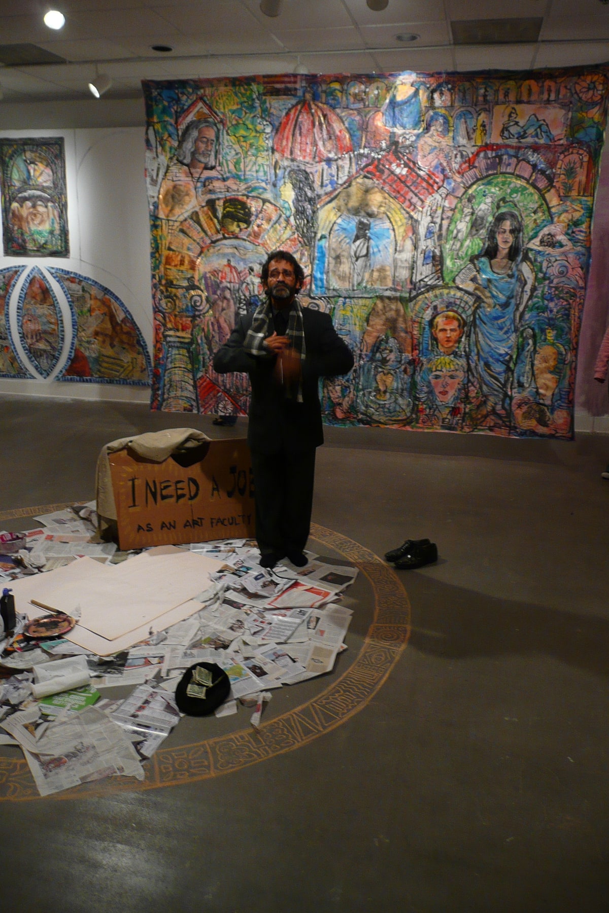 An artist stands among painted canvases, newspapers and drawing materials in a gallery installation.