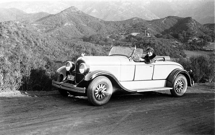Donald A. Hall Sr.'s 1925 Studebaker Roadster parked on an unpaved mountain road with a smiling woman behind the wheel against layered hills.