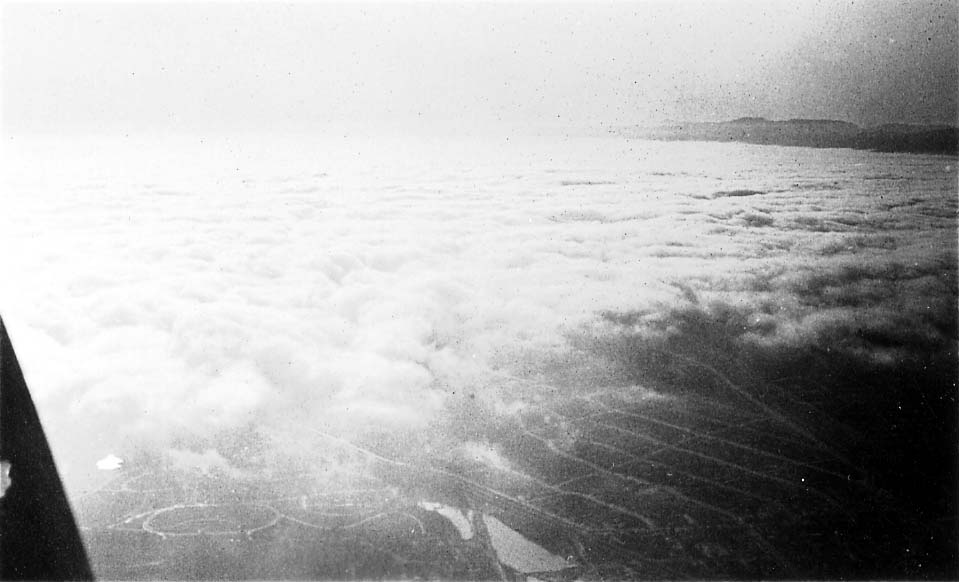 An aerial view looking past an aircraft strut at a thick blanket of white clouds, with a patch of coastal land visible below.