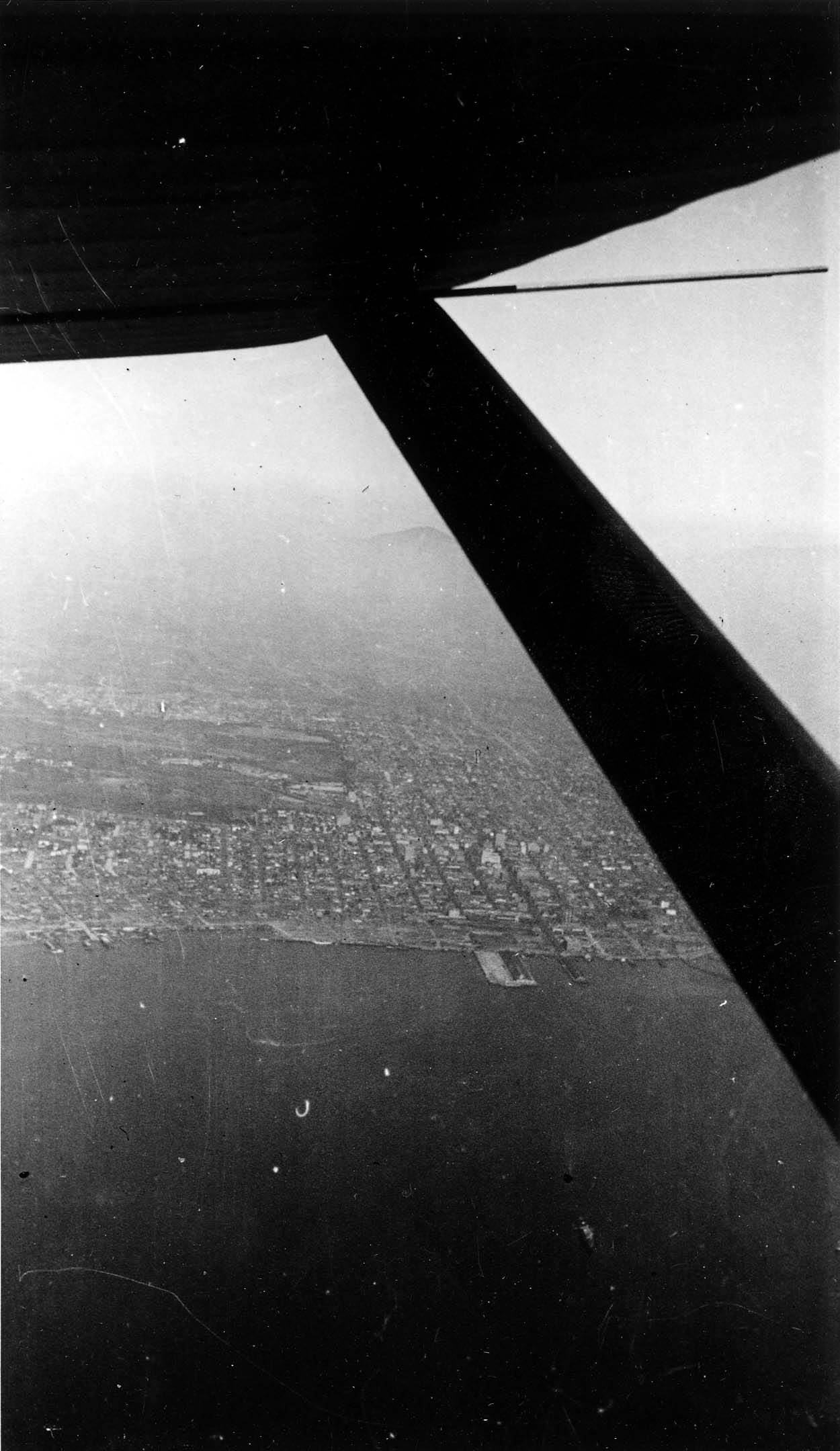 An aerial view framed by the underside of an aircraft wing, looking down at a coastal city grid meeting the ocean.