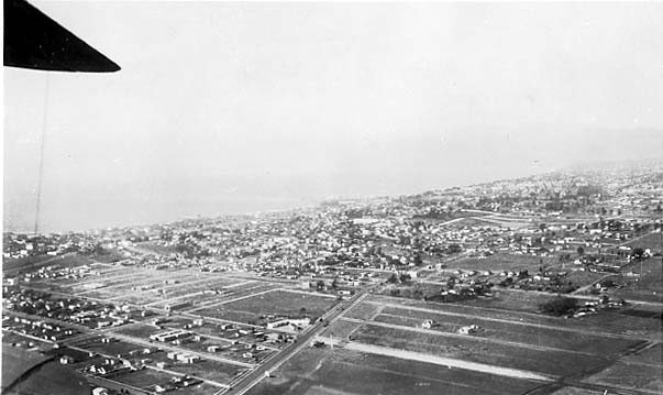 An aerial view looking out from under an aircraft wing toward a sprawling coastal city grid bordering the Pacific ocean.