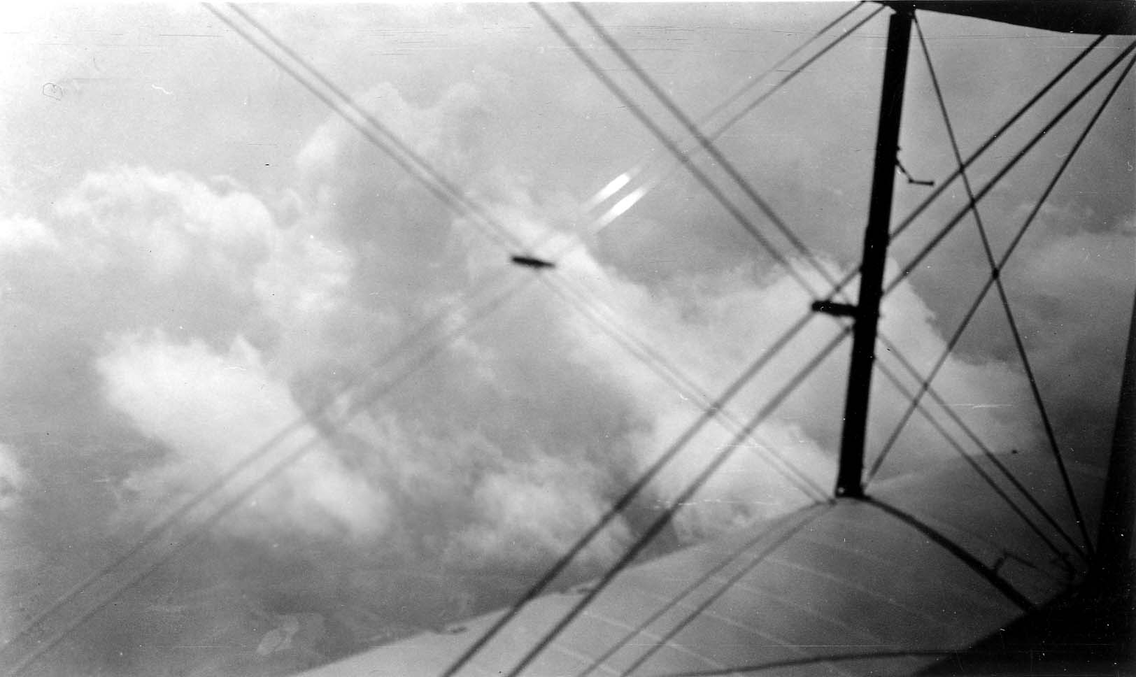 An aerial view looking through the struts and tension wires of a biplane at thick, puffy white clouds.