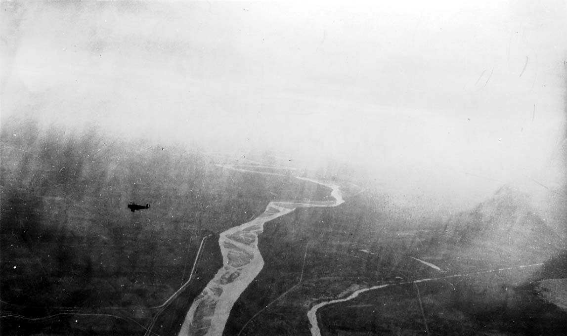 An aerial view of a winding river valley shrouded in mist, with a small silhouette of another airplane flying in the distance.