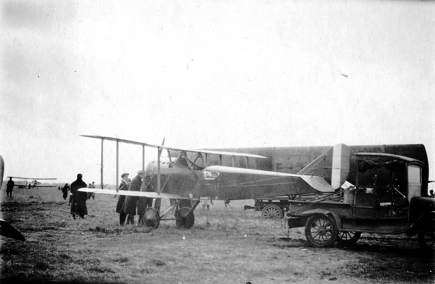 A vintage cylindrical tanker truck parked beside a military biplane on a grass airfield as ground crew mechanics perform maintenance.