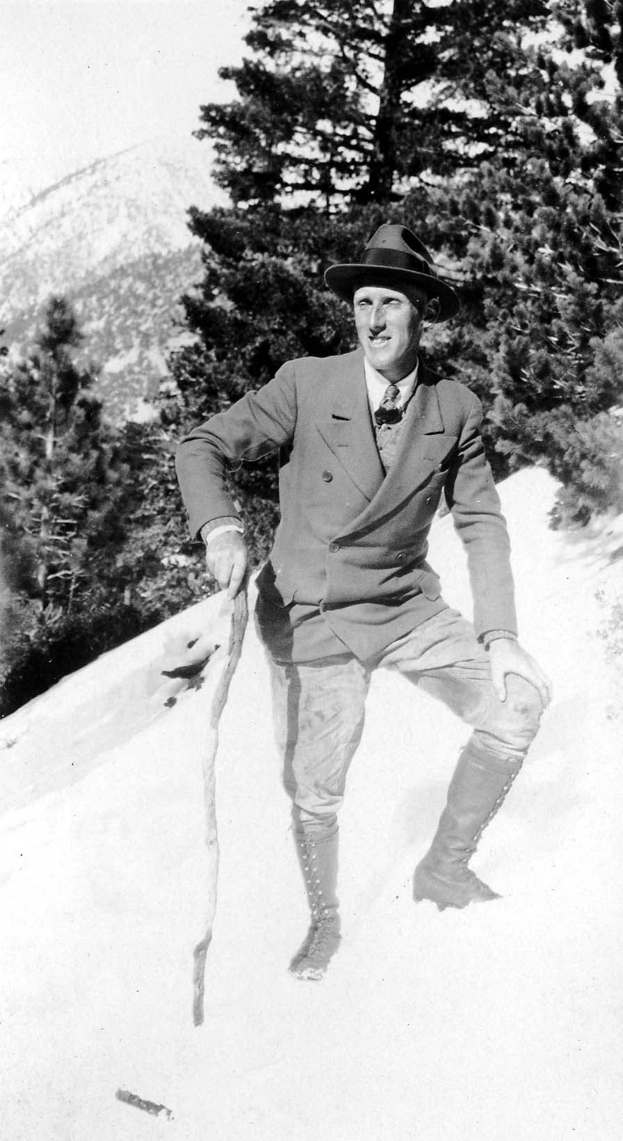 A smiling hiker in a fedora, double-breasted jacket, breeches, and high lace-up boots holding a rustic walking stick on a snowy mountain incline.