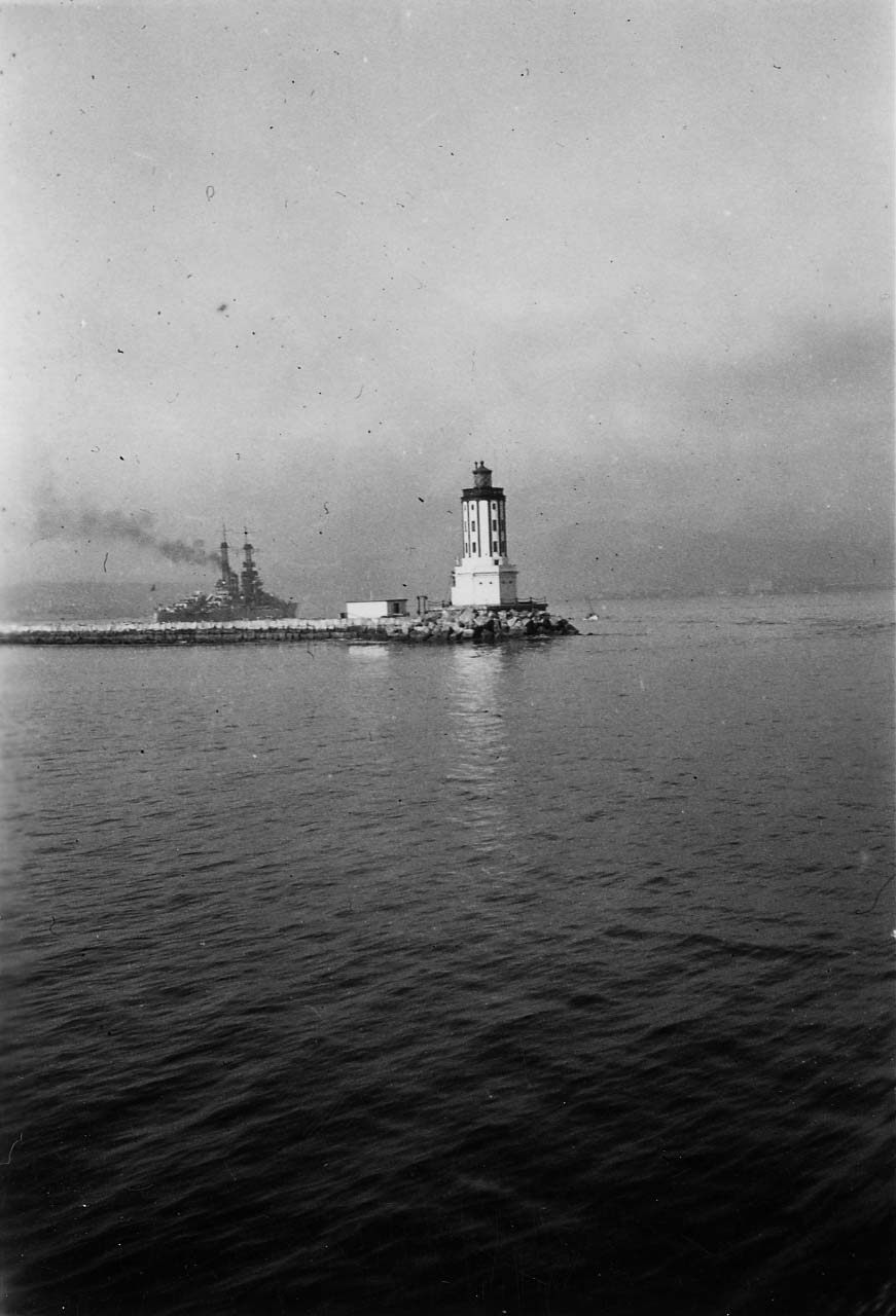A view across Los Angeles Harbor toward the historic Angels Gate Lighthouse at the end of the breakwater, with a U.S. Navy cage-mast battleship steaming in the distance.
