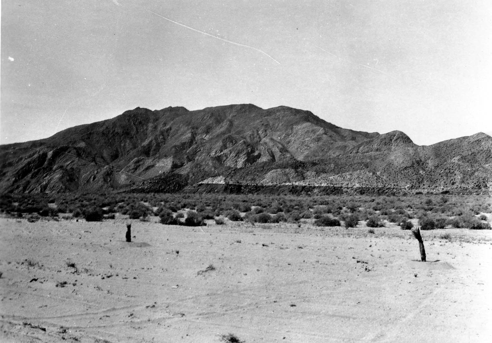 A flat, barren desert plain with sparse low-lying brush, leading up to a massive, rugged mountain range under a clear sky.