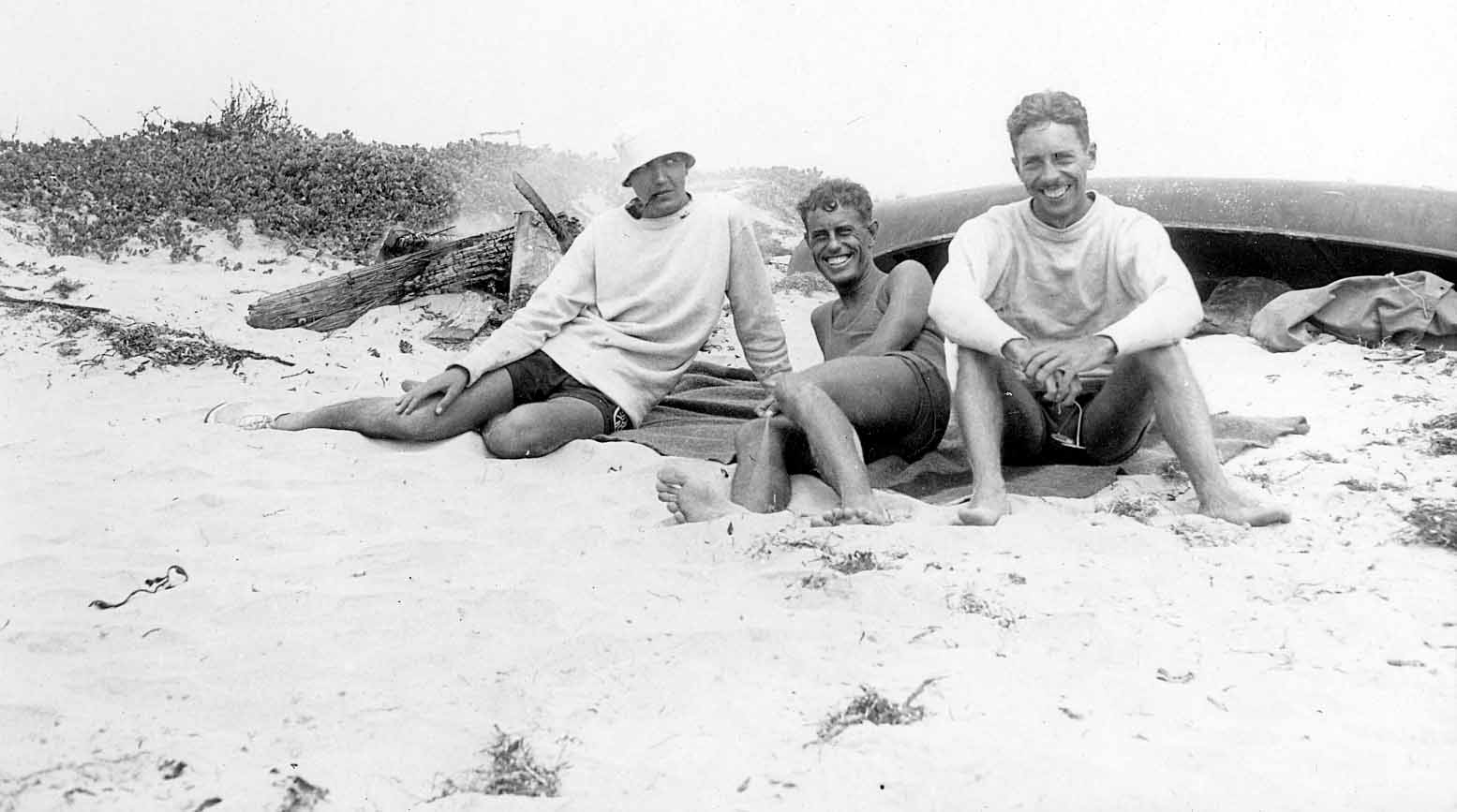 Donald A. Hall Sr. and two friends sitting on the beach sand next to a dark canoe.