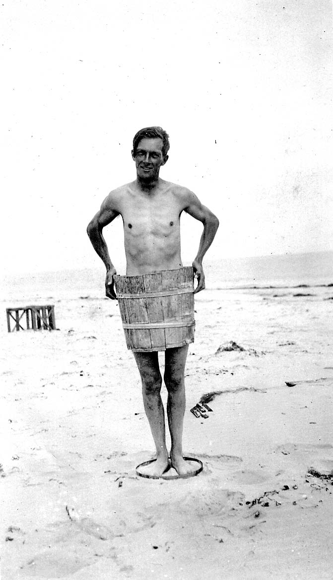 A smiling young man standing on a sandy beach wearing a wooden barrel around his waist and standing inside a metal hoop as a humorous beach gag.