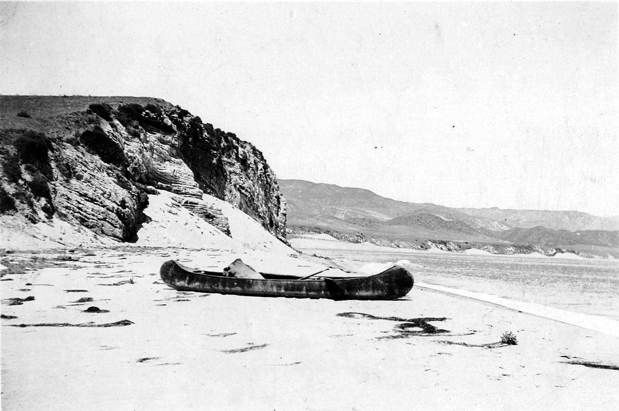 Donald A. Hall Sr.'s wooden canoe resting on a sandy beach at the base of towering stratified ocean bluffs, with calm sea water in the background.