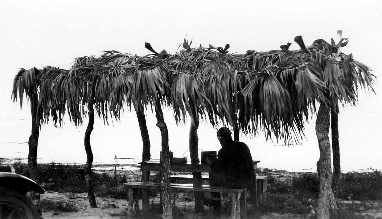A silhouette of a person sitting at a rustic wooden bench under a thatched palm frond ramada on a sandy beach, beside the fender of a parked vintage automobile.