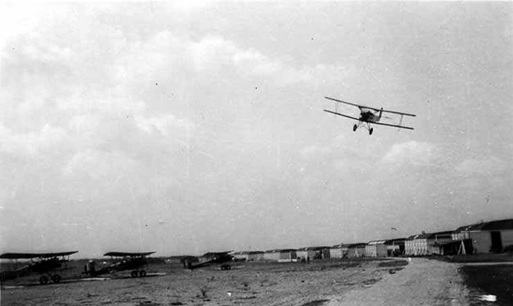 A high-quality action photograph of a training biplane climbing into the sky after takeoff, flying over the long line of hangars at Brooks Field.