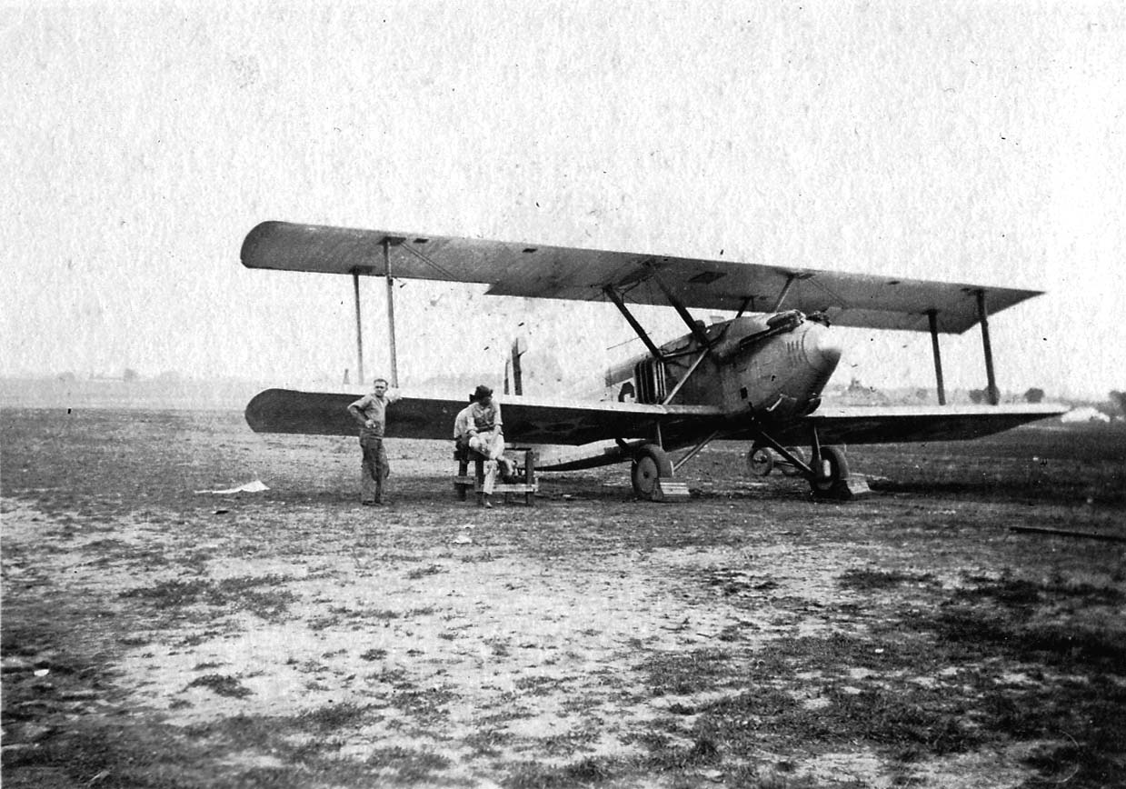 Ground crew mechanics resting on a tool chest beneath the lower wing of an experimental pursuit biplane with a streamlined nose spinner and tunnel radiator.
