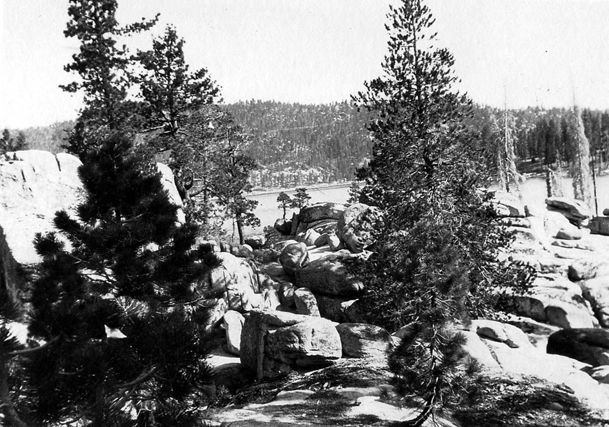 Granitic boulders and young pine trees clustered along the rocky shoreline of a mountain lake.