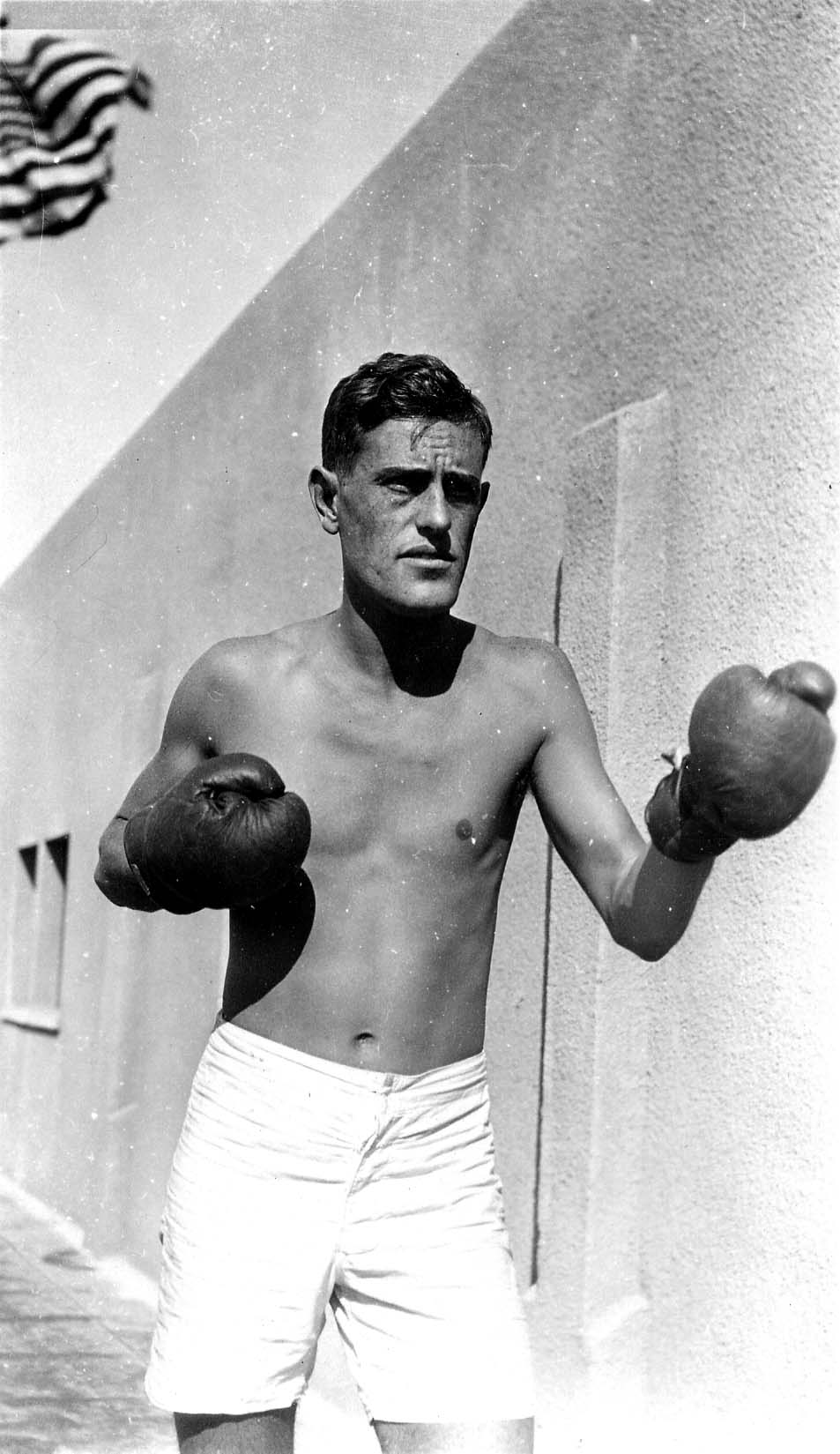 A shirtless Donald A. Hall Sr. wearing boxing gloves, standing in an outdoor fighting stance next to a building with an American flag.