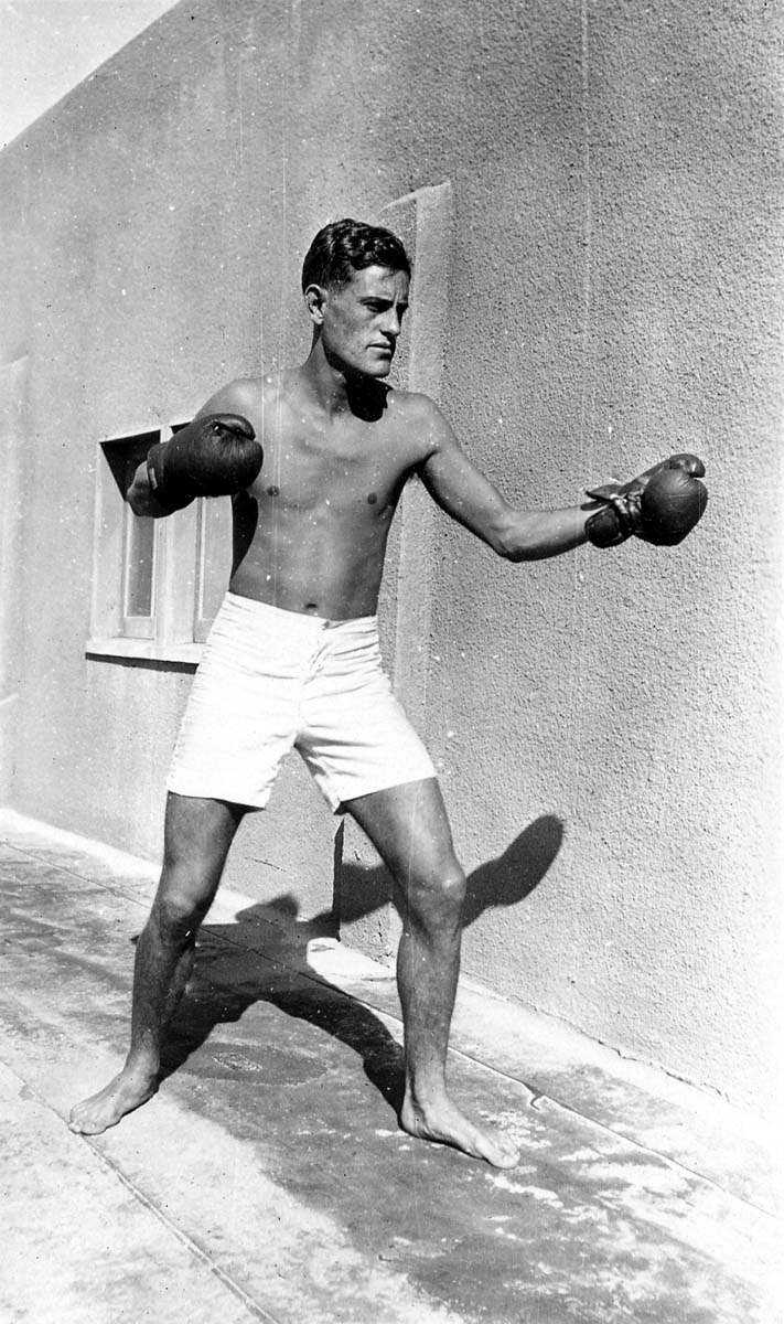A shirtless Donald A. Hall Sr. in white trunks and boxing gloves, posing in a fighting stance casting a sharp shadow against a stucco wall.
