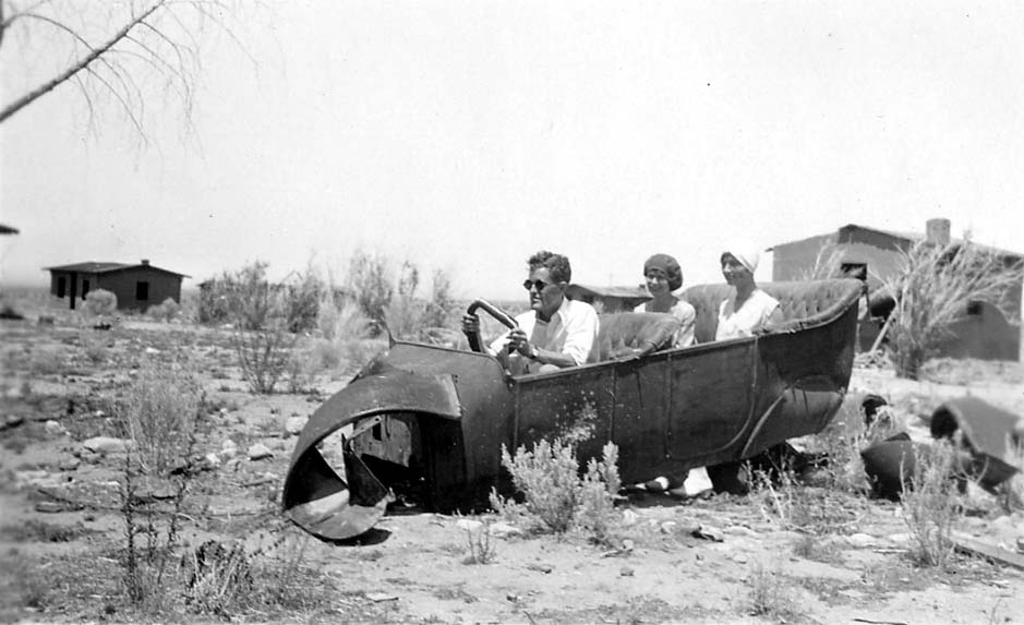 Donald A. Hall Sr. wearing dark sunglasses and pretending to drive a broken, hollowed-out car shell in the desert alongside two women.