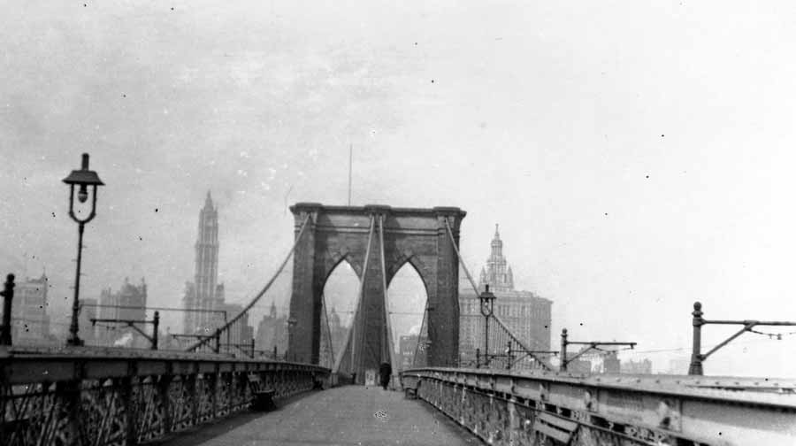 A wide perspective looking down the wooden pedestrian promenade of the Brooklyn Bridge, framing its massive Gothic stone arches and suspension cables against the Manhattan skyline.