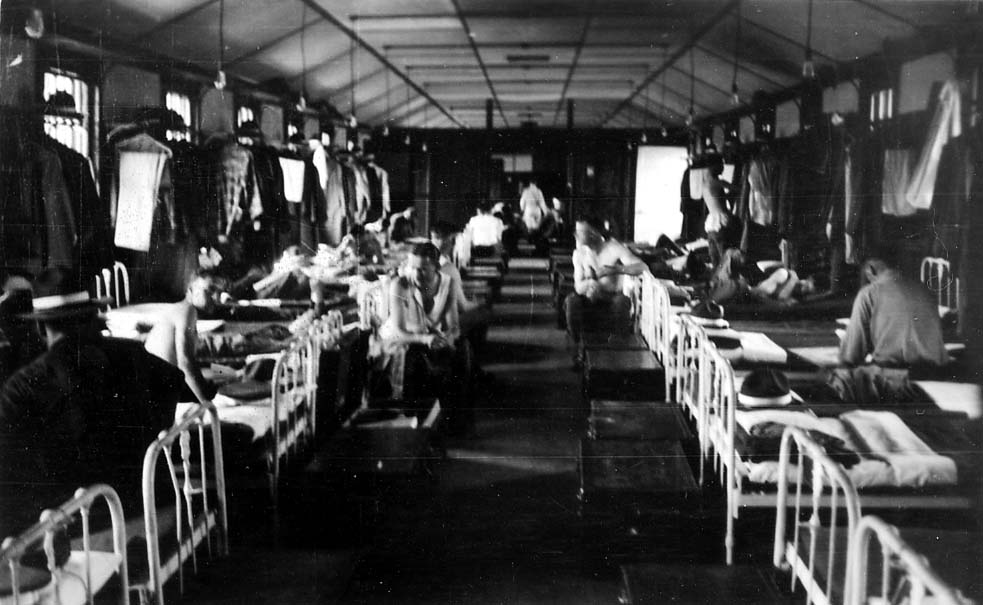 A wide interior view of the wooden Brooks Field military barracks, showing flight cadets relaxing on narrow iron cots with uniforms hanging from the rafters.