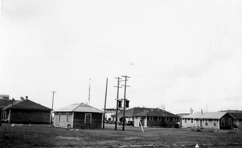 A view of wooden military barracks and buildings at Brooks Field, Texas, featuring utility poles and a small airplane model mounted on a high pole.