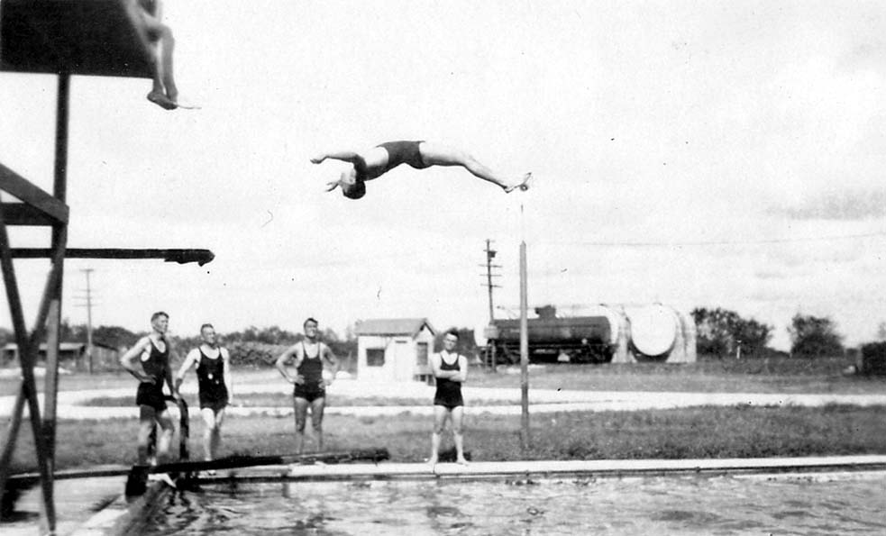 An action shot of a flight cadet executing a mid-air backflip into a swimming pool, while a group of fellow cadets watches from the deck, with barracks and rail tank cars in the background.