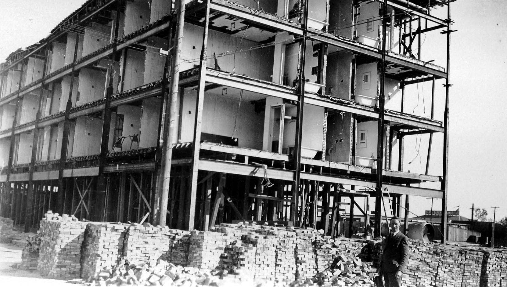 A multi-story building under construction, showing the bare steel I-beam framework and partially completed interior walls, with stacks of bricks in the foreground.