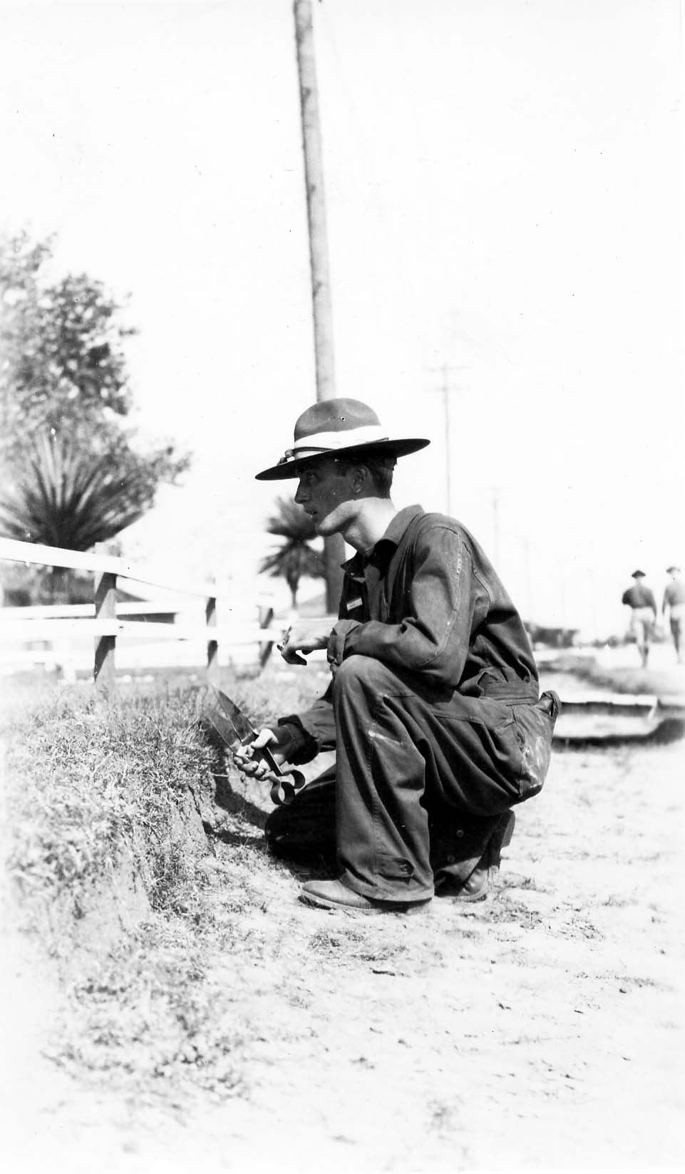 A flight cadet in coveralls crouching on the ground, using large hand clippers to manually trim the grass next to a white fence.