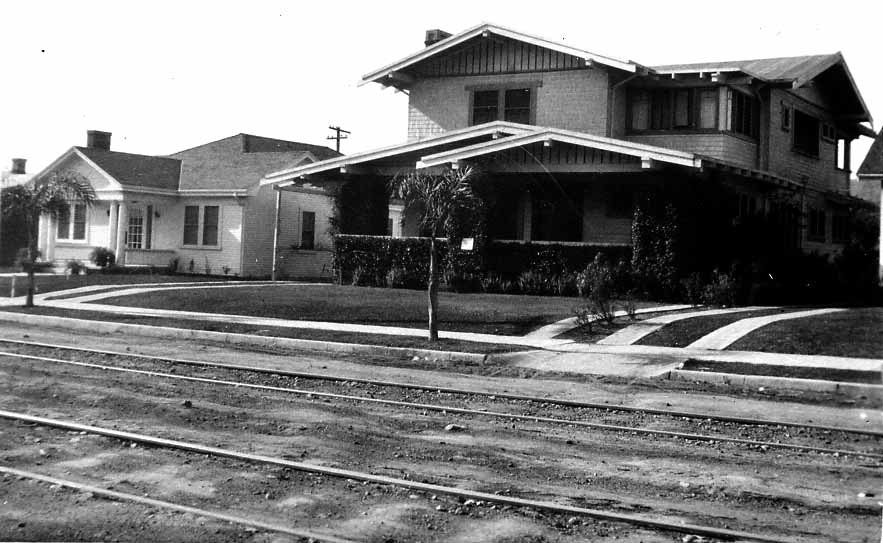 A two-story California Craftsman airplane bungalow with deep gabled eaves, wide front porch, and unpaved streetcar tracks in the foreground.