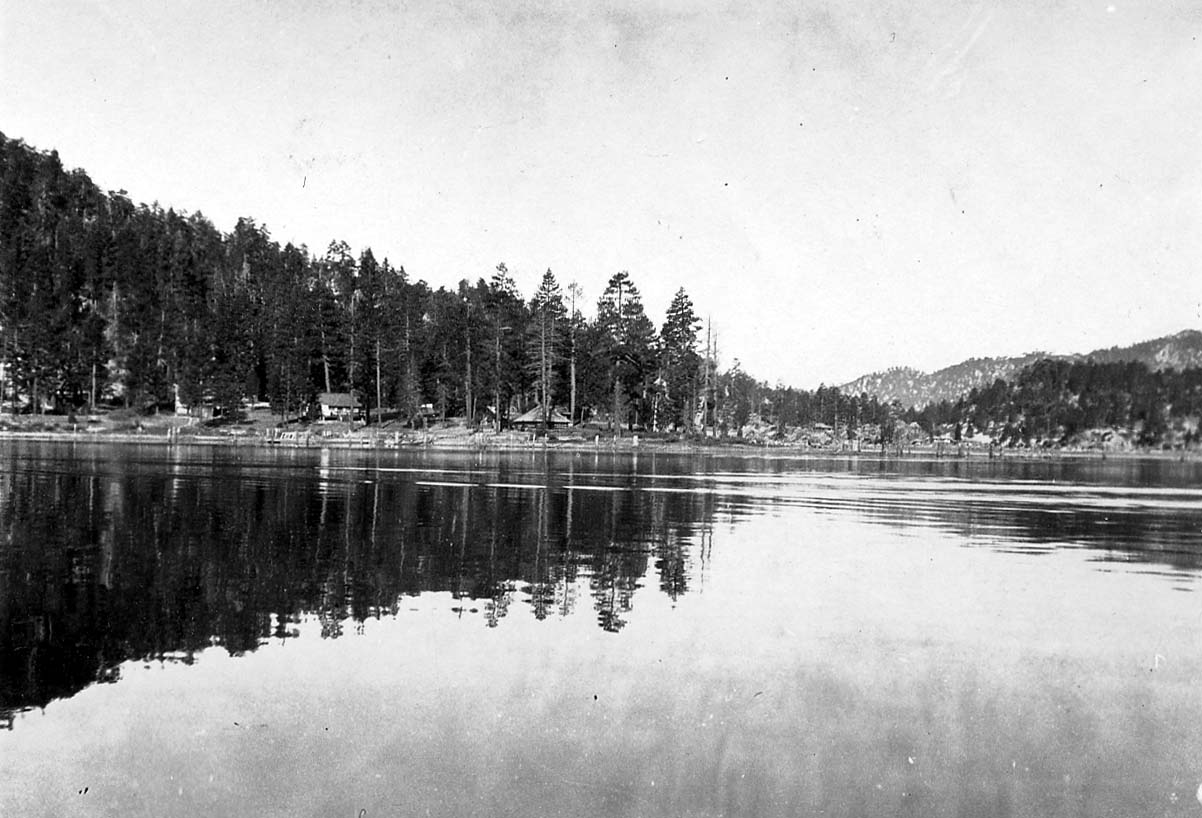A calm mountain lake reflecting a dense forest and rustic lakeside cabins along the opposite tree-lined shore.