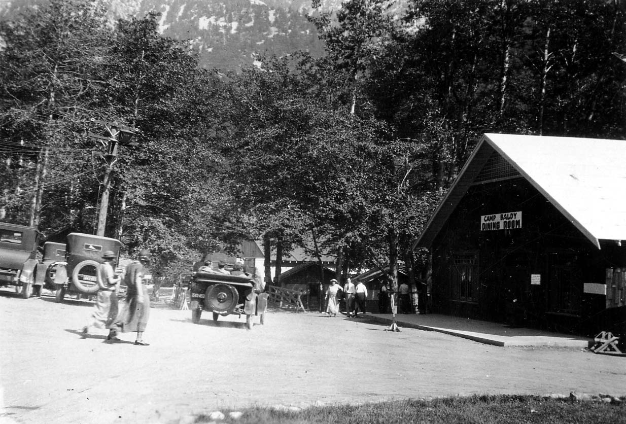 A rustic wooden building with a sign reading "CAMP BALDY DINING ROOM" nestled among pine trees, with vintage cars parked nearby.