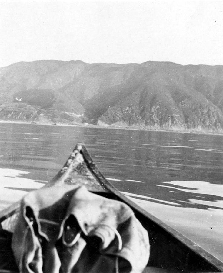 A view over the wooden bow of a canoe floating on calm ocean water, looking toward steep, rugged mountain slopes rising directly from the shoreline.