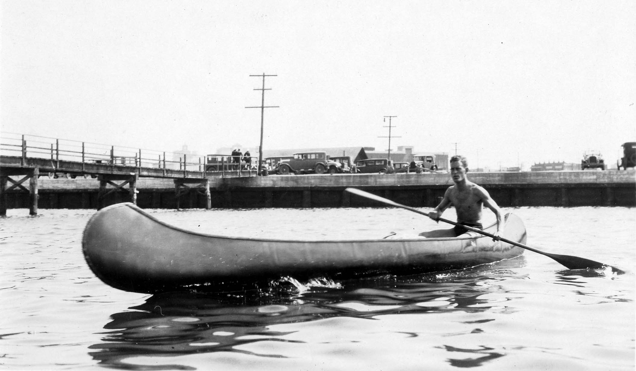 A shirtless Donald A. Hall Sr. paddling his handcrafted canoe across calm coastal waters with a wooden pier and parked cars behind him.