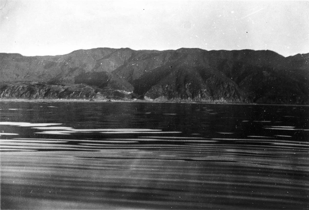 Calm ocean waters showing smooth glassy slick bands and gentle surface swells reflecting beneath steep coastal mountain slopes.