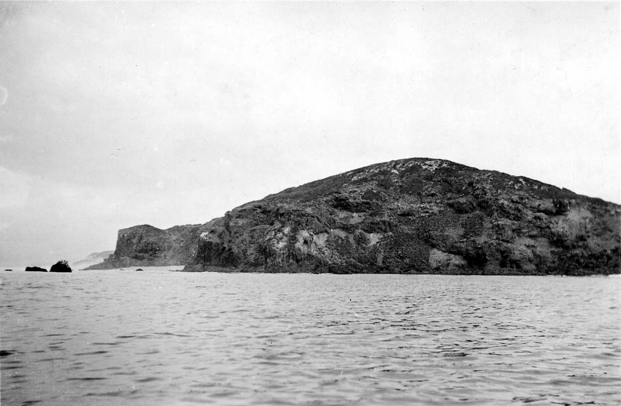 A rugged, volcanic coastal headland with steep rocky cliffs and small offshore sea stacks rising from a gentle ocean swell.