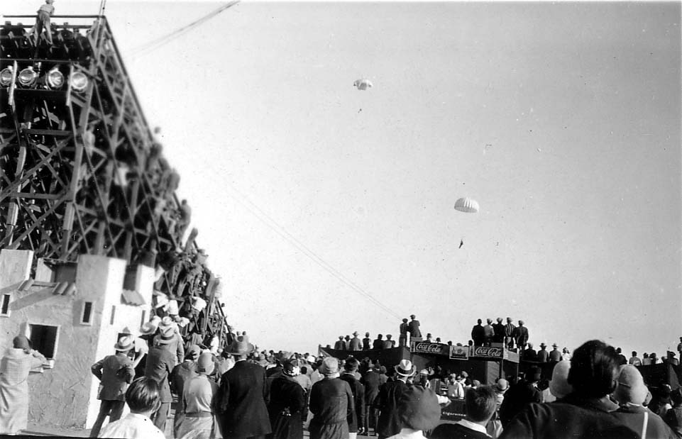 A crowd of people watching two parachute jumpers descending from the sky above a tall wooden scaffolding structure.