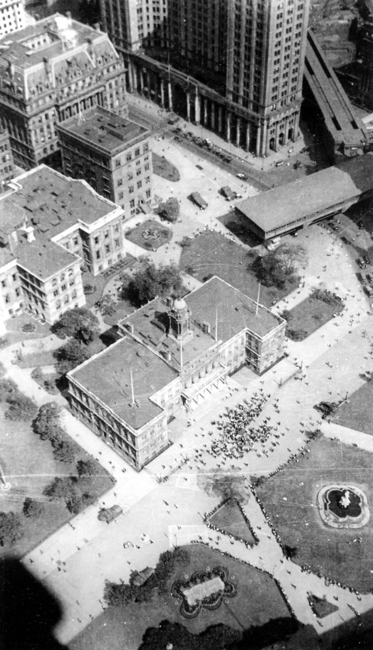 A high-angle downward perspective from the Woolworth Building over City Hall Park, showing a massive crowd gathered in front of City Hall for the Lindbergh reception.