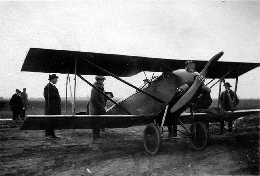 Three men in suits and coats inspecting a small, high-speed single-seat racing biplane with a wooden propeller on an unpaved airfield.