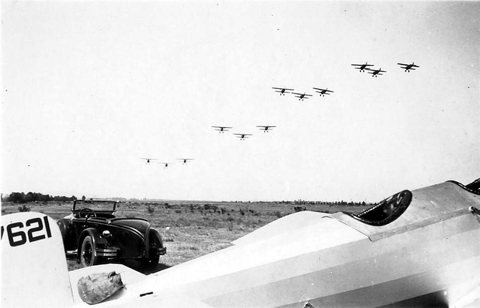 The completed Hall X-1 tandem-wing prototype parked on an airfield beside a roadster as an eight-plane military biplane formation flies overhead.