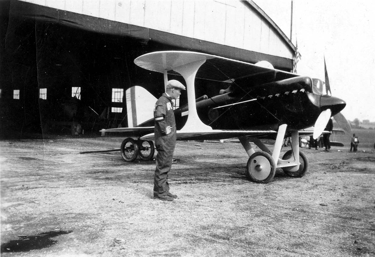 A sleek black Curtiss R2C-1 racing biplane with streamlined I-struts and Curtiss-Reed metal propeller parked outside an airfield hangar beside a mechanic.