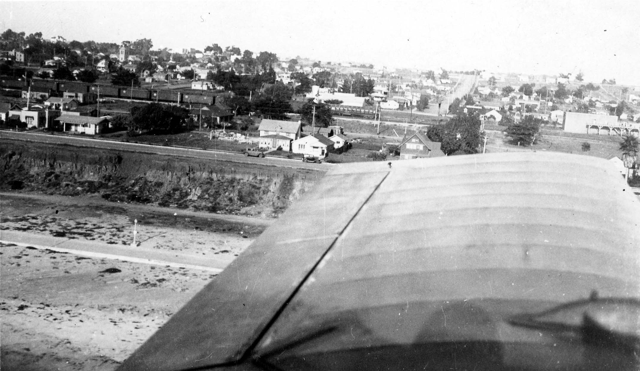 An aerial view framed past an aircraft wing showing a coastal Southern California town, beach bluffs, and freight trains on the tracks.