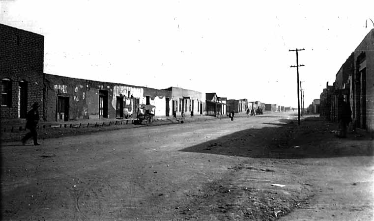A wide view looking down a wide, unpaved dirt street lined with flat-roofed adobe buildings and wooden porticos in a sun-baked desert town.