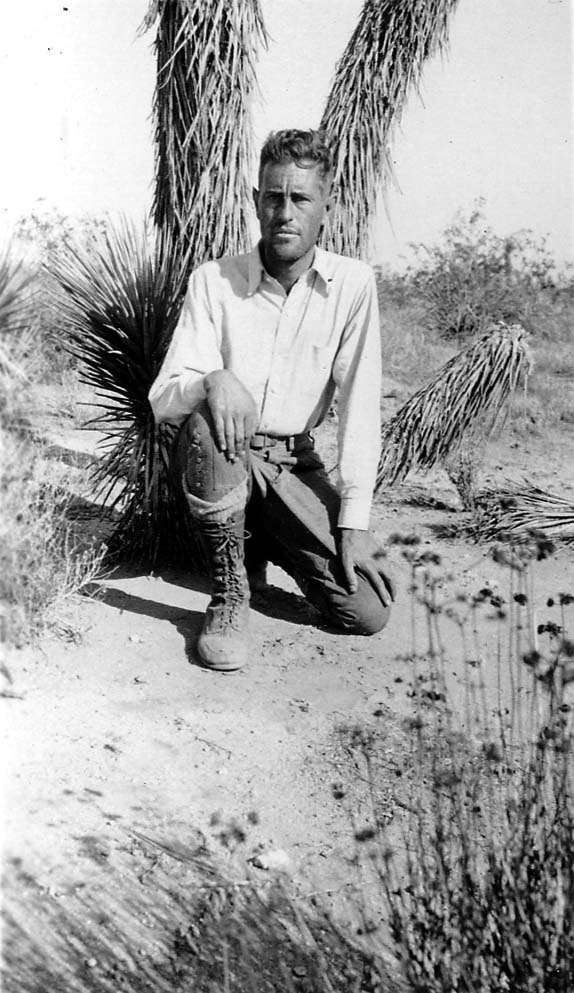 Donald A. Hall Sr. kneeling in the desert sand in front of yucca plants, wearing a light shirt and tall lace-up hiking boots.