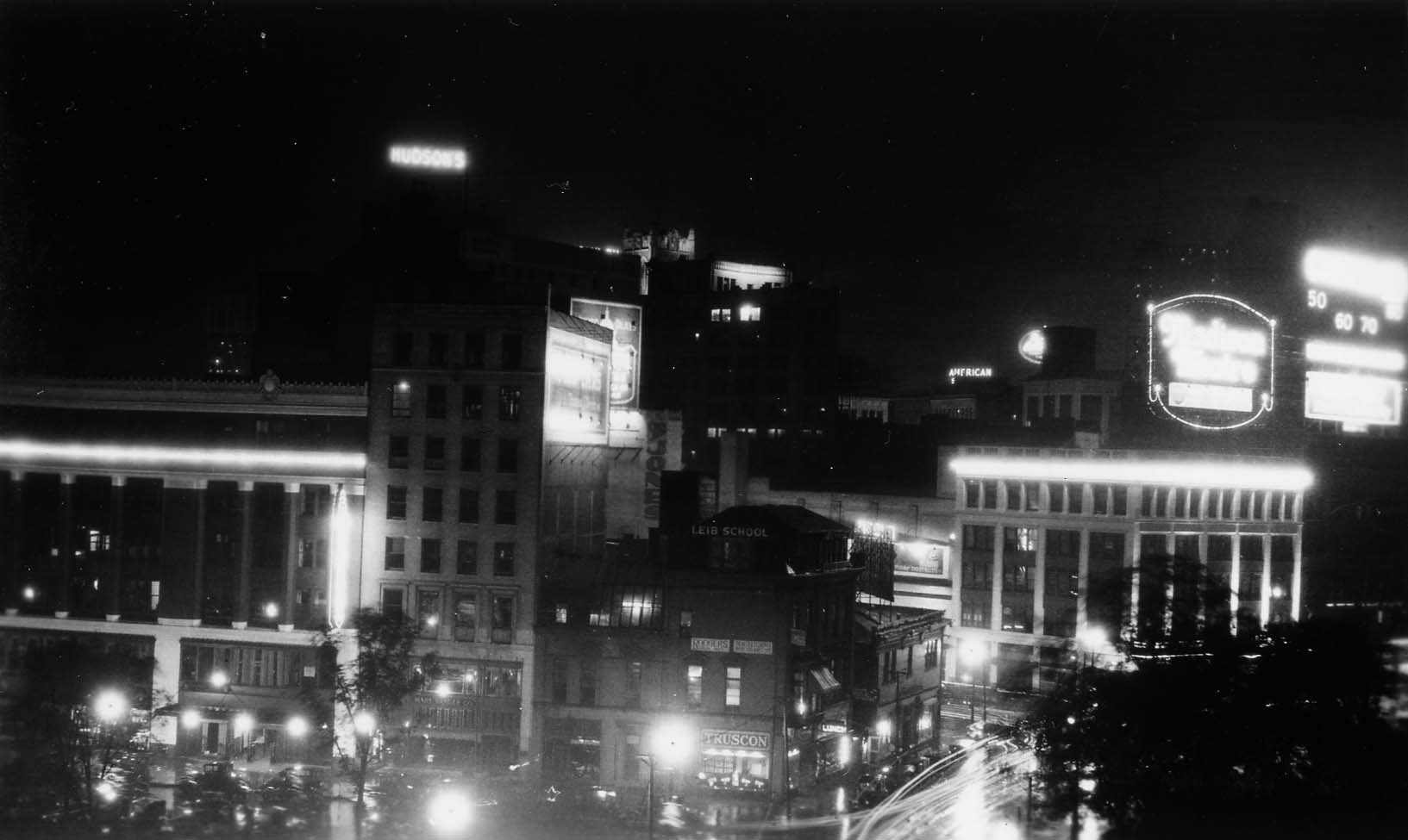 An elevated view over electrified commercial buildings in Downtown Detroit, featuring an illuminated rooftop sign for Hudson's department store and light trails from streetcars.