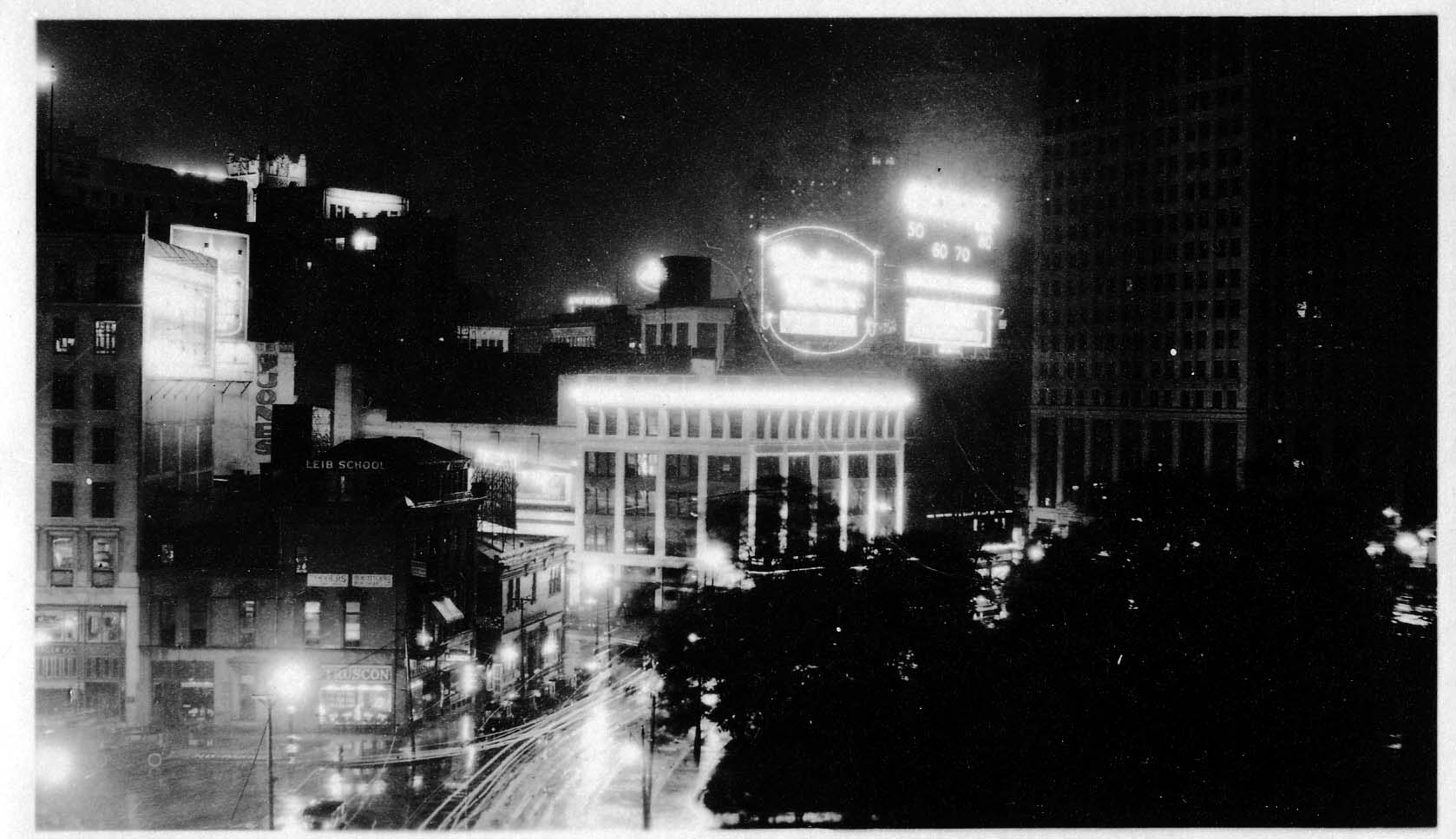 A wide-angle night photograph across Grand Circus Park toward the glowing Madison Theatre sign, illuminated commercial blocks, and the David Whitney Building under a dark sky.