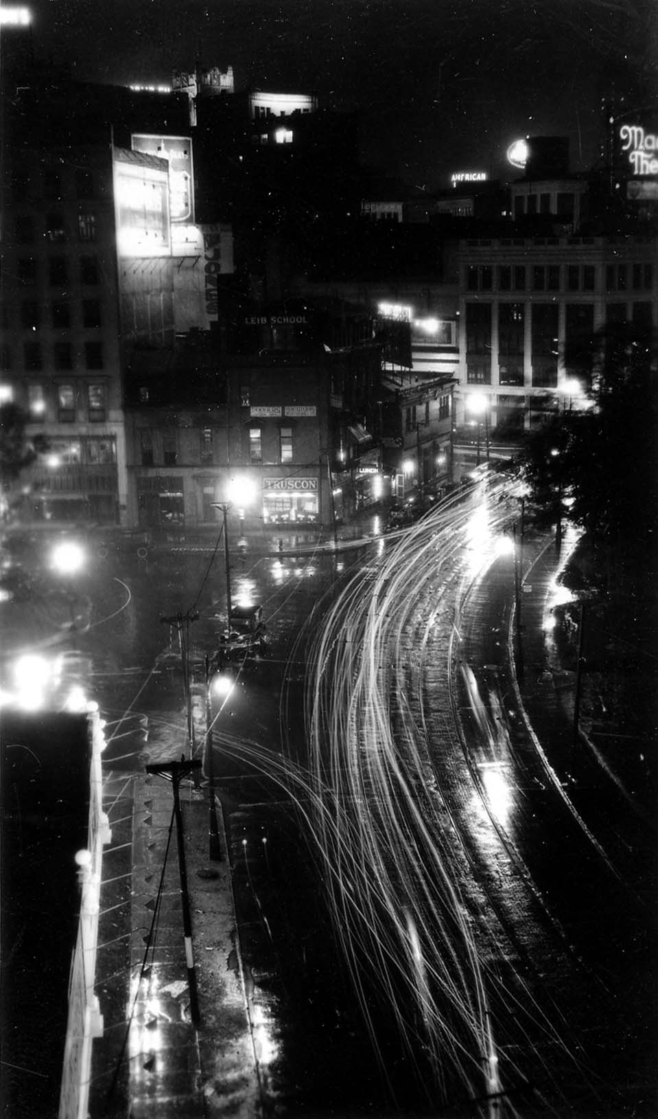 A vertical long-exposure looking down onto rain-soaked Grand Circus Park streets, capturing dense white headlight trails sweeping past multi-story commercial buildings and glowing storefronts.