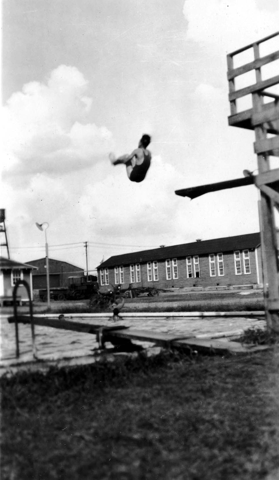 An action shot of a man mid-dive off a wooden springboard into a community swimming pool, with brick buildings and a wooden tower in the background.