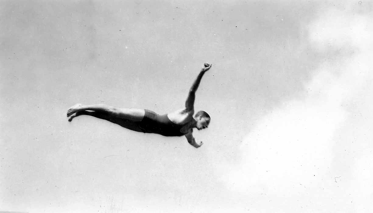 A high-contrast photograph of a man in a swimsuit executing a swan dive, frozen in mid-air against a bright, cloudy sky.