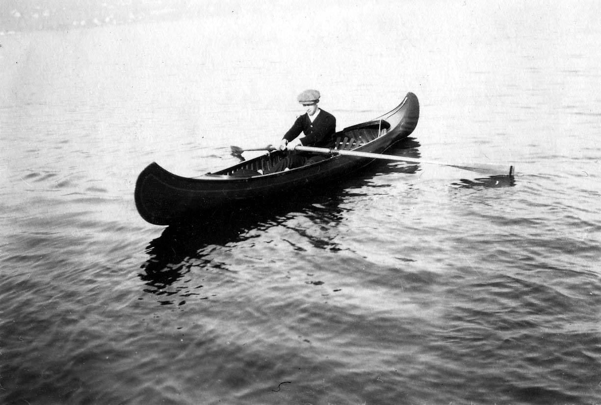 Donald A. Hall Sr. wearing a flat cap and sweater, sitting in a canoe and rowing with long wooden oars.
