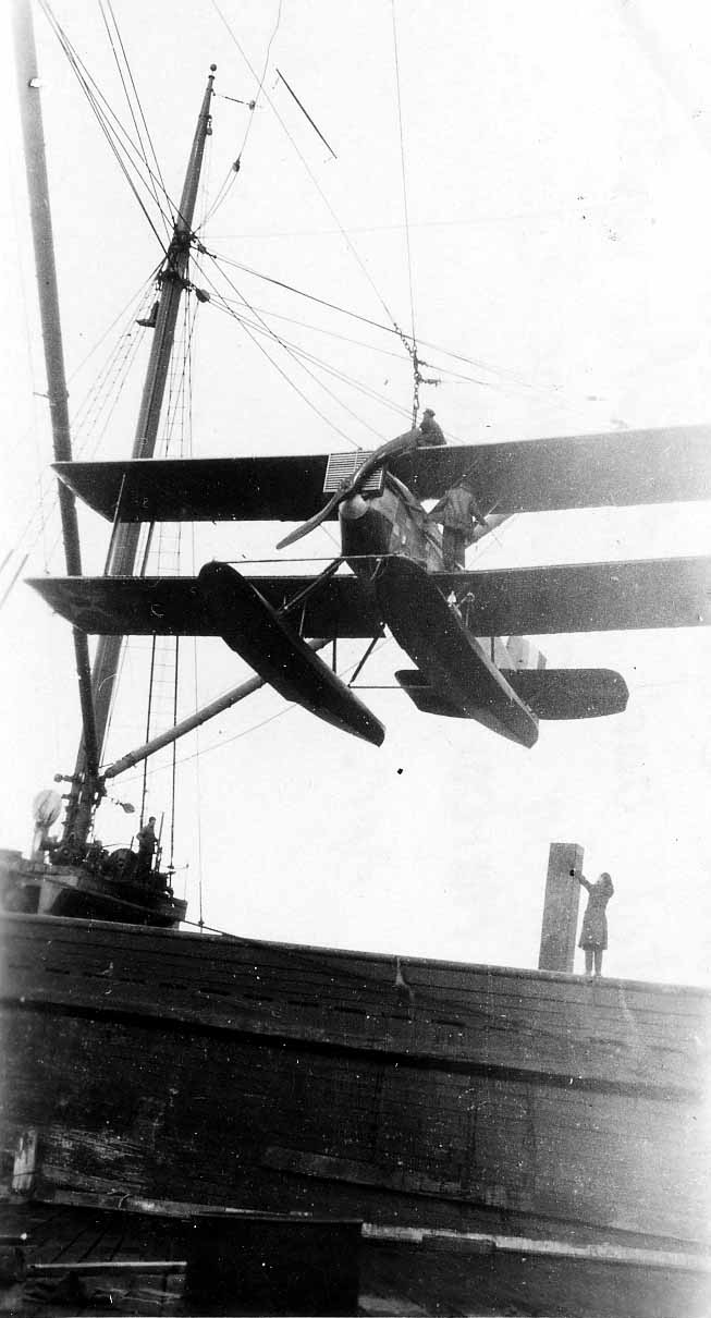 A full profile view of a Douglas World Cruiser seaplane dangling in mid-air from a ship's derrick alongside the tall wooden mast.