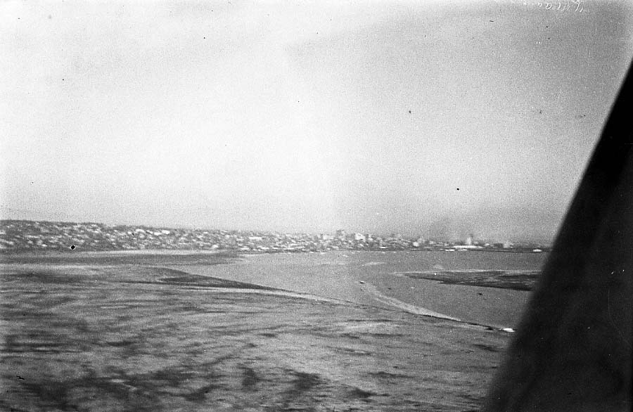 An aerial view looking out past an aircraft wing towards a coastline and city, showing the Dutch Flats area where a plane may be taking off.